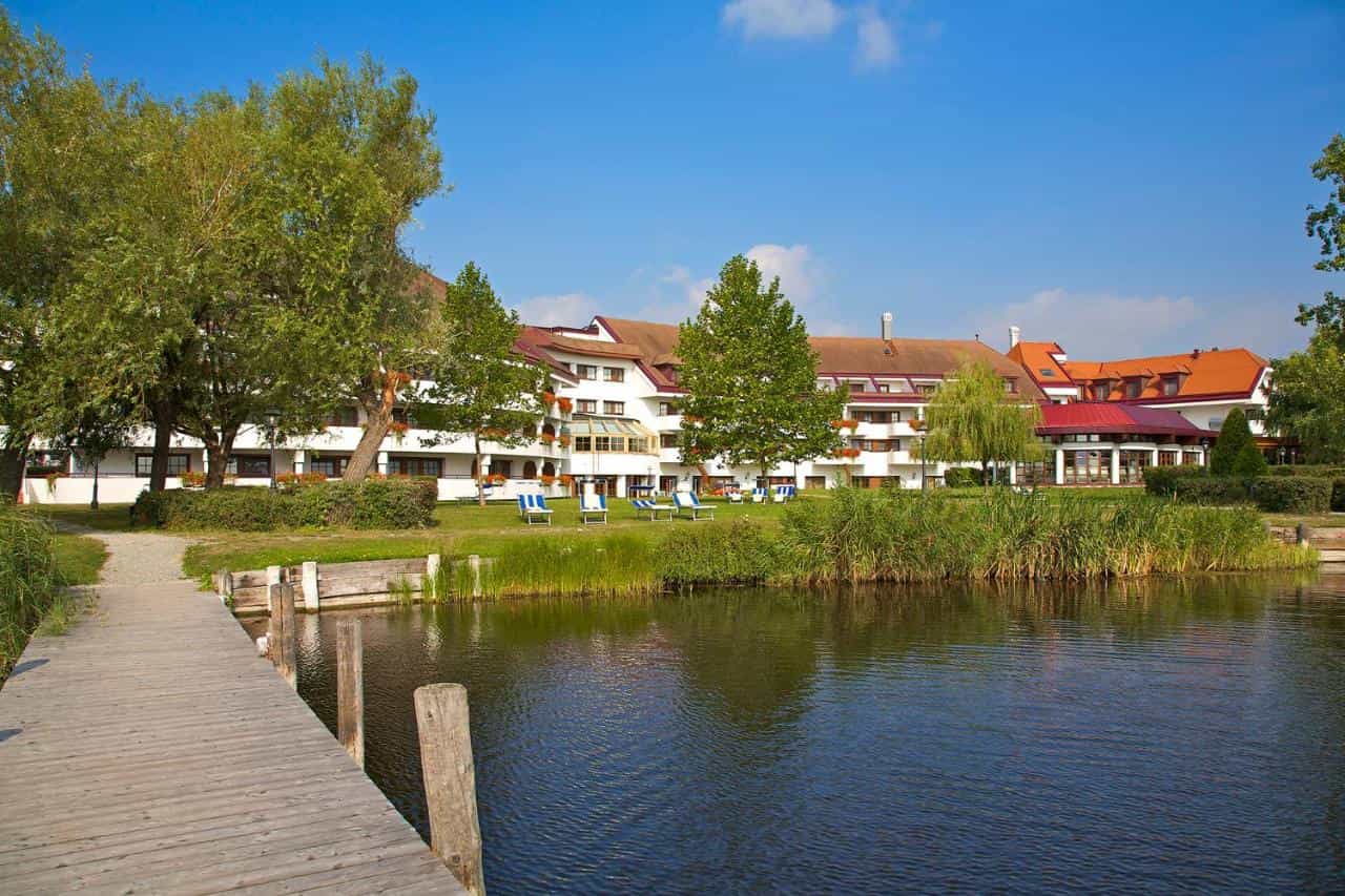 Seehotel Rust on lake with wooden dock and green lawn chairs under blue sky.