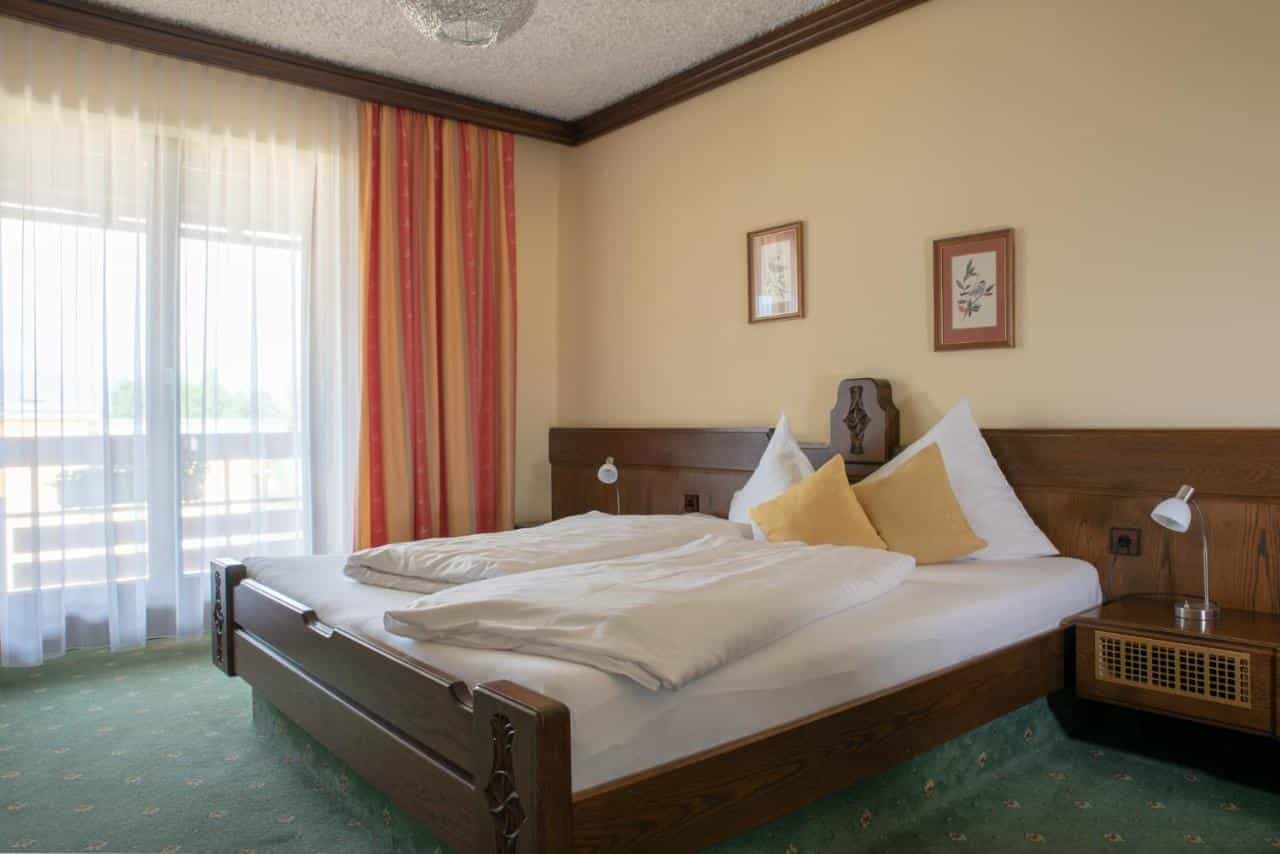 Hotel room with wooden bed, white duvets, yellow pillows, and balcony access through sheer curtains.