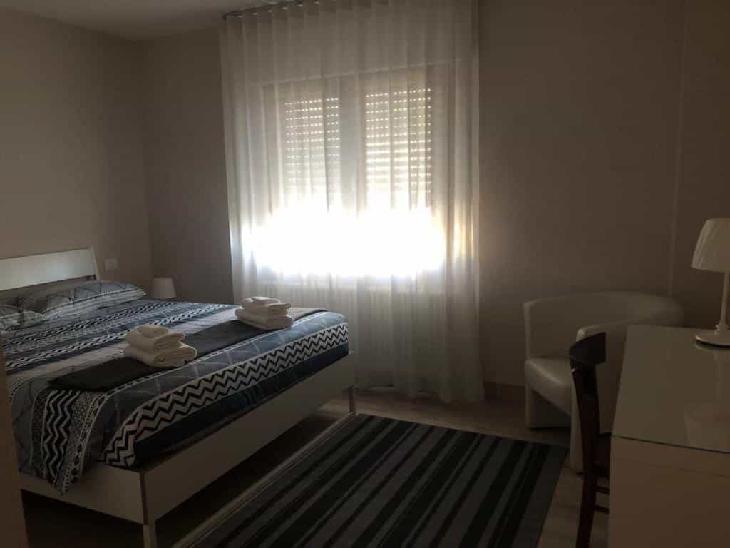 Bedroom with blue patterned bedding, white armchair, desk, and bright window with sheer curtains.