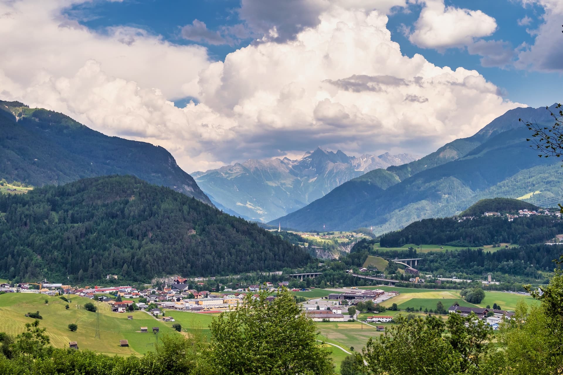 Town of Imst in Tirol valley with green hills, distant alpine peaks, and dramatic clouds.