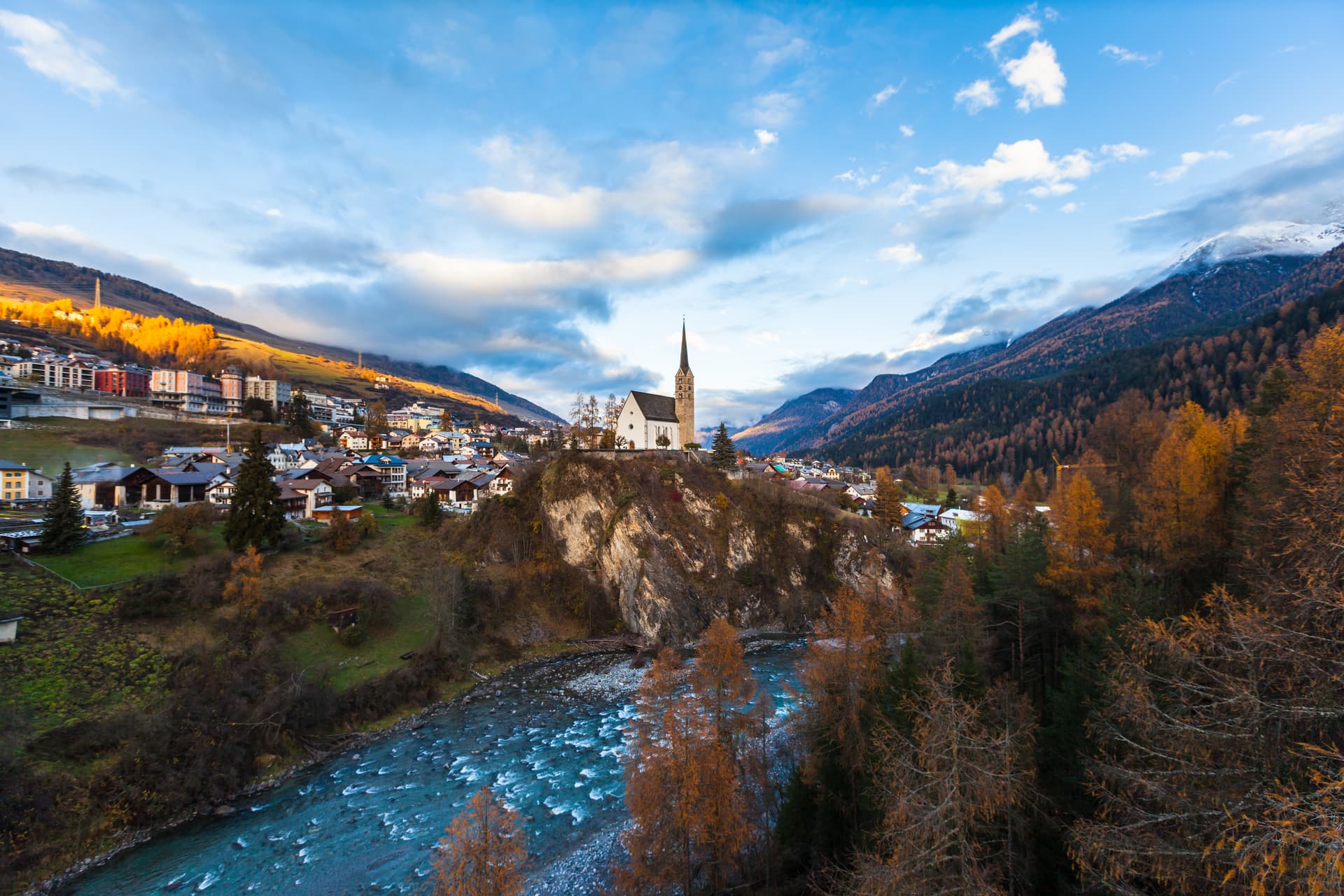 Alpine town of Scuol in sunset with church on cliff above rushing river and autumn mountains.