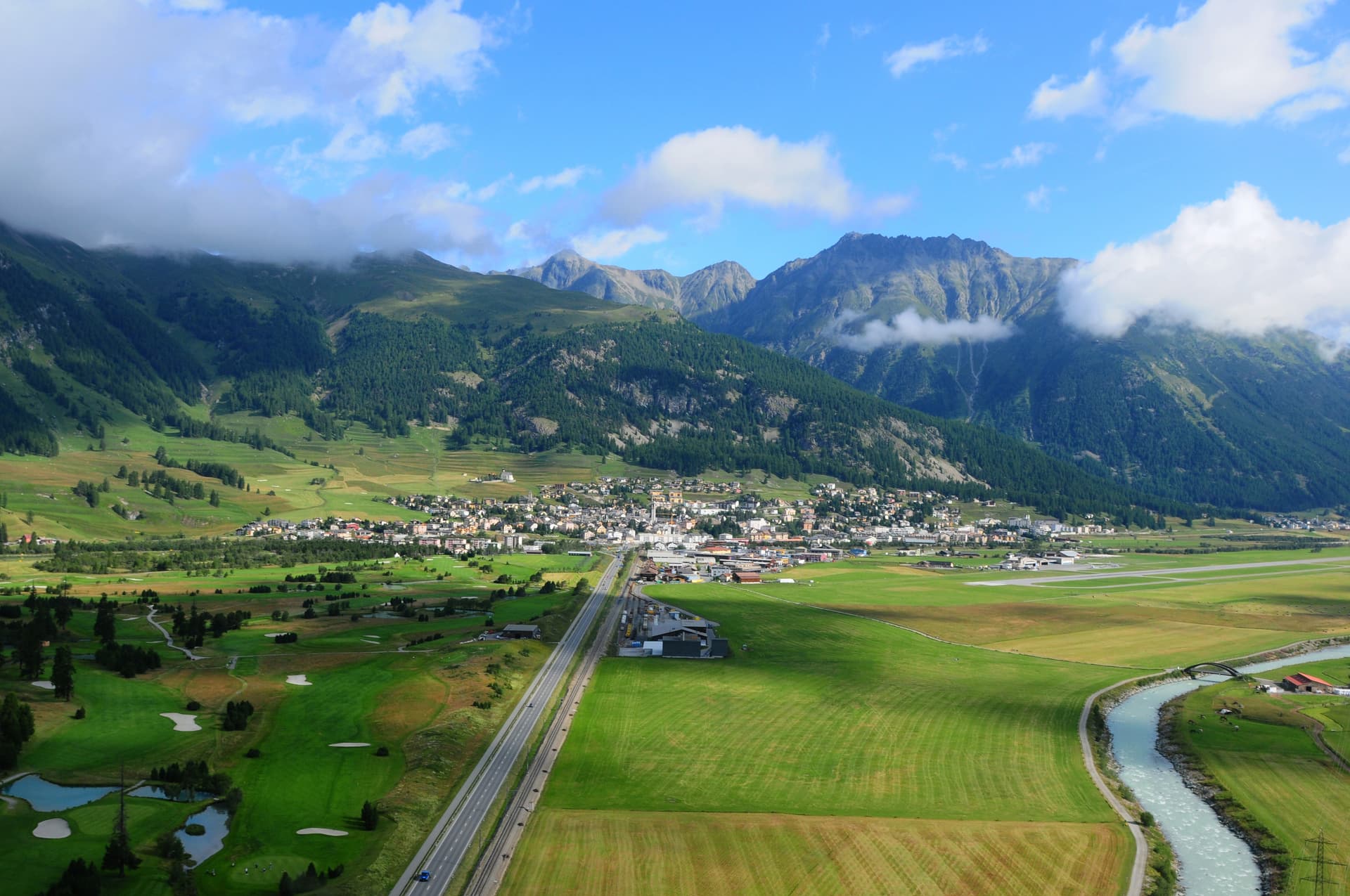 Aerial view of Samedan Airport, town, green valley, river, and high alpine mountains.