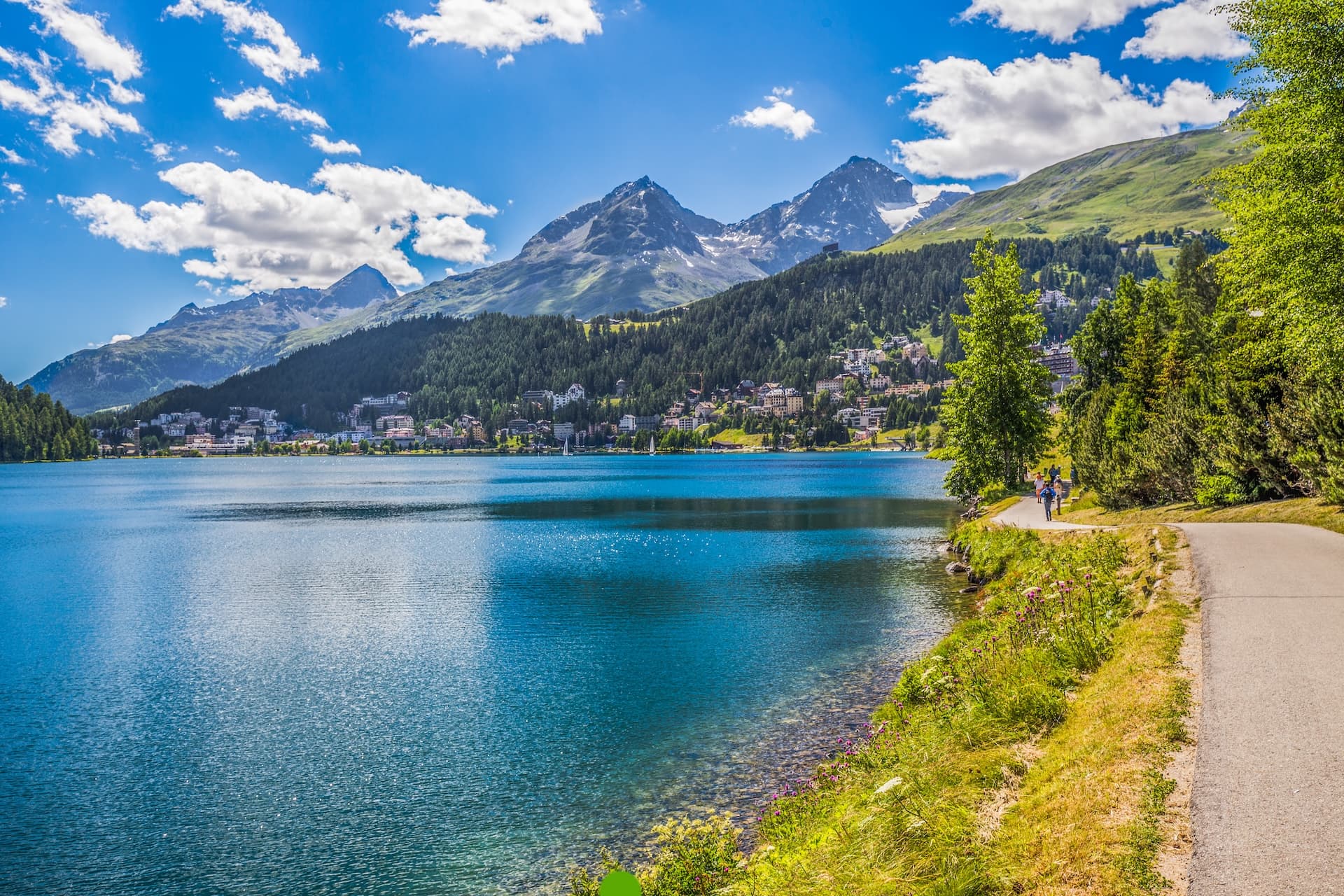 People walking along path beside Lake St. Moritz with Swiss Alps and town in background