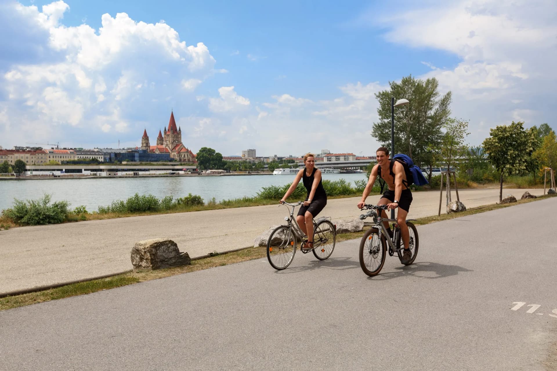 Two people cycling on paved path by river with distinctive red-roofed church in distance