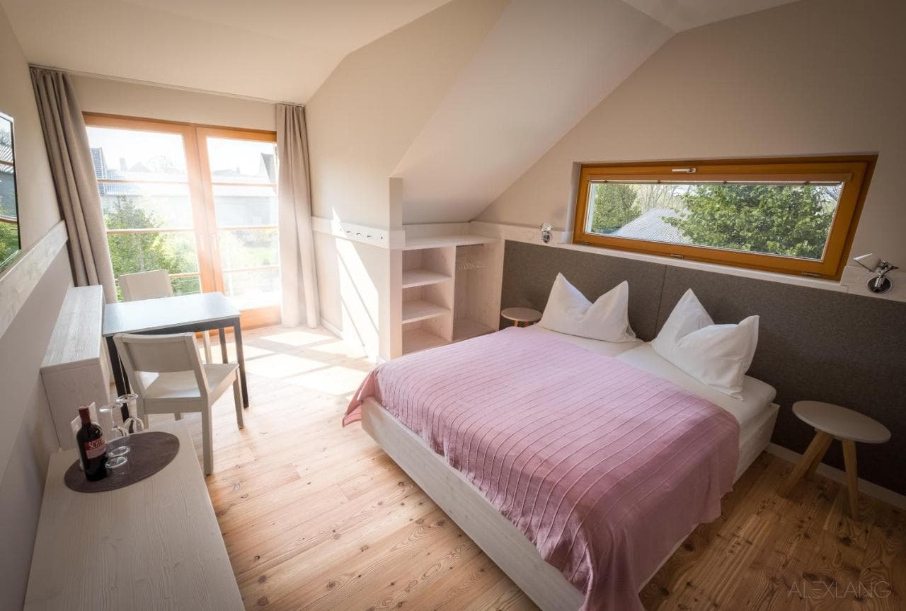 Attic guest room with wooden floors, pink bedspread, and bright window overlooking greenery at SCHEIBER-Wein-&-Gästehaus.