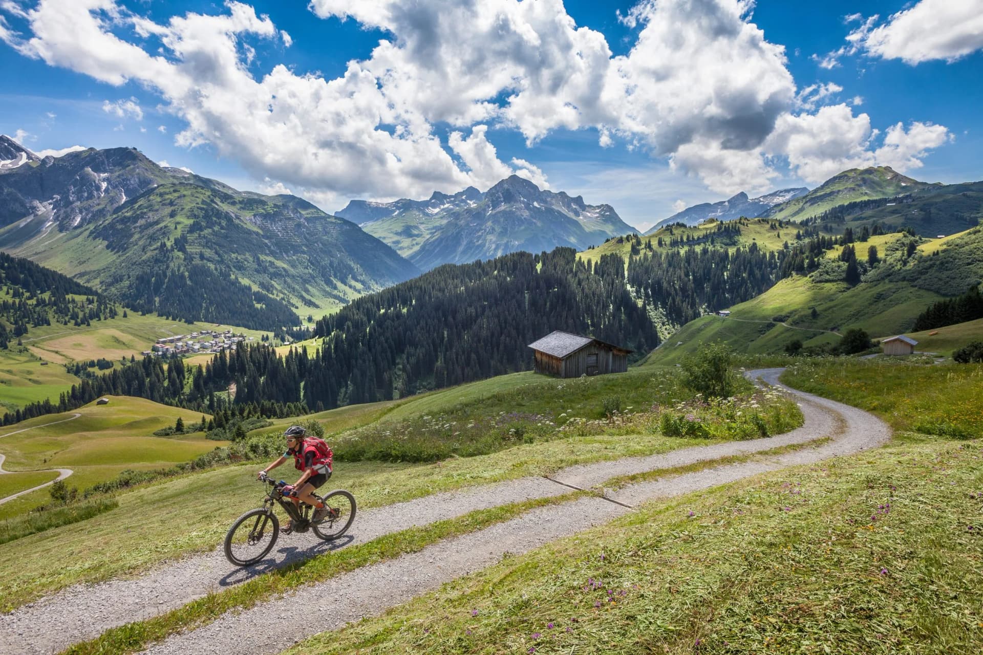 Mountain biking on gravel path through green alpine meadows with village and snowy peaks.
