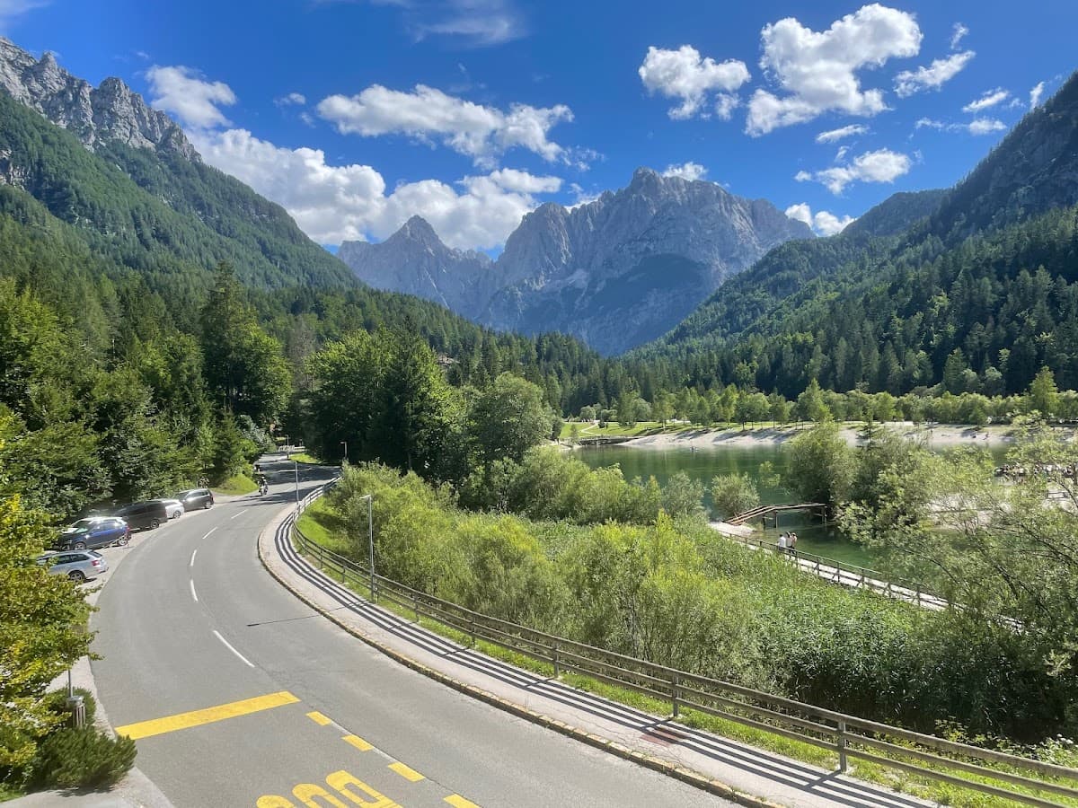 Winding road near a lake surrounded by lush green mountains under a blue sky with clouds.