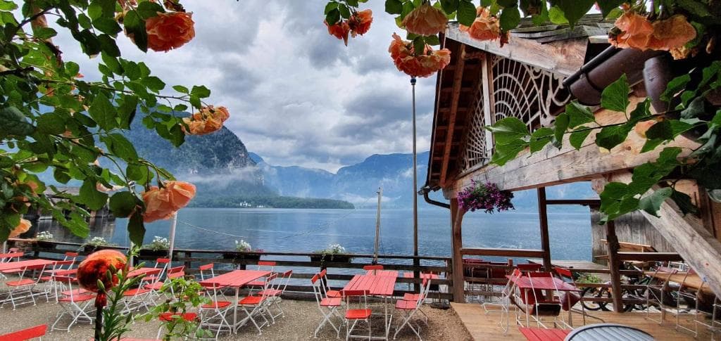 Outdoor cafe with red tables overlooking a mountain lake and forest under a cloudy sky.