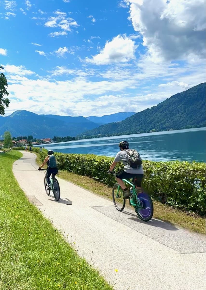 Cyclists riding along a paved path beside a lake with mountains in the background