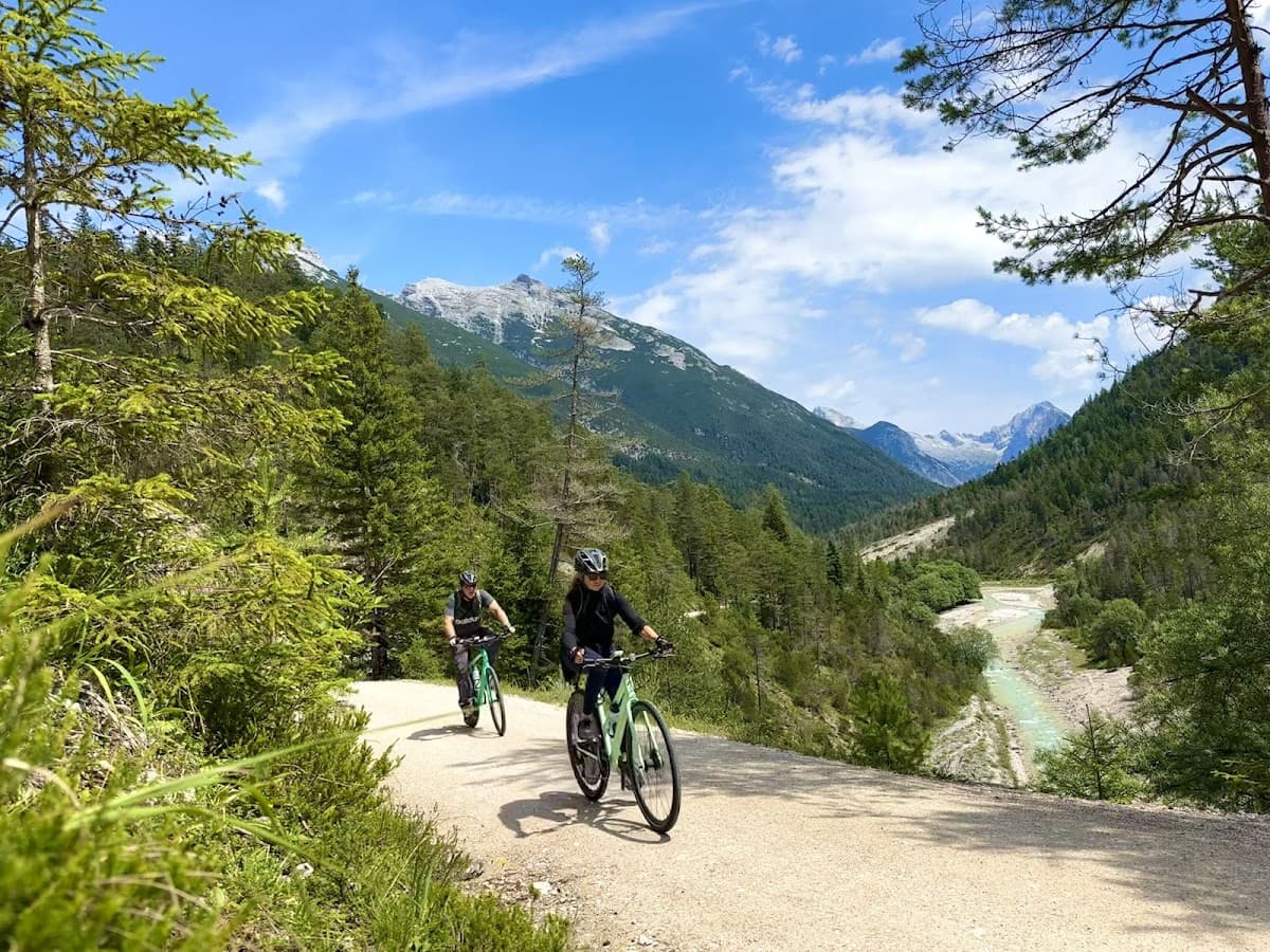 Cyclists riding mountain bikes on a gravel path through an alpine valley with pine trees and river.