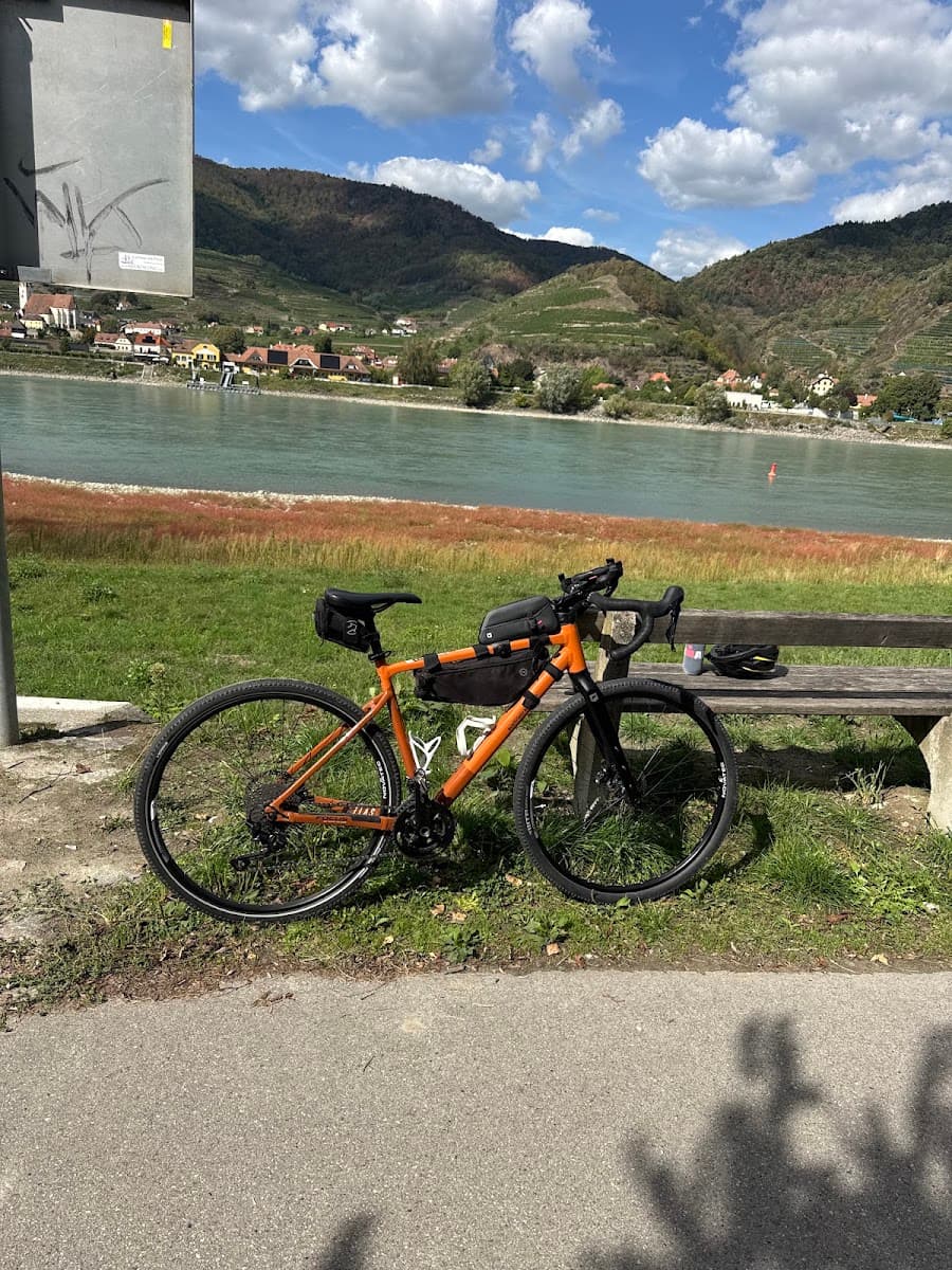 Orange gravel bicycle parked near a river with hills and a village in the background