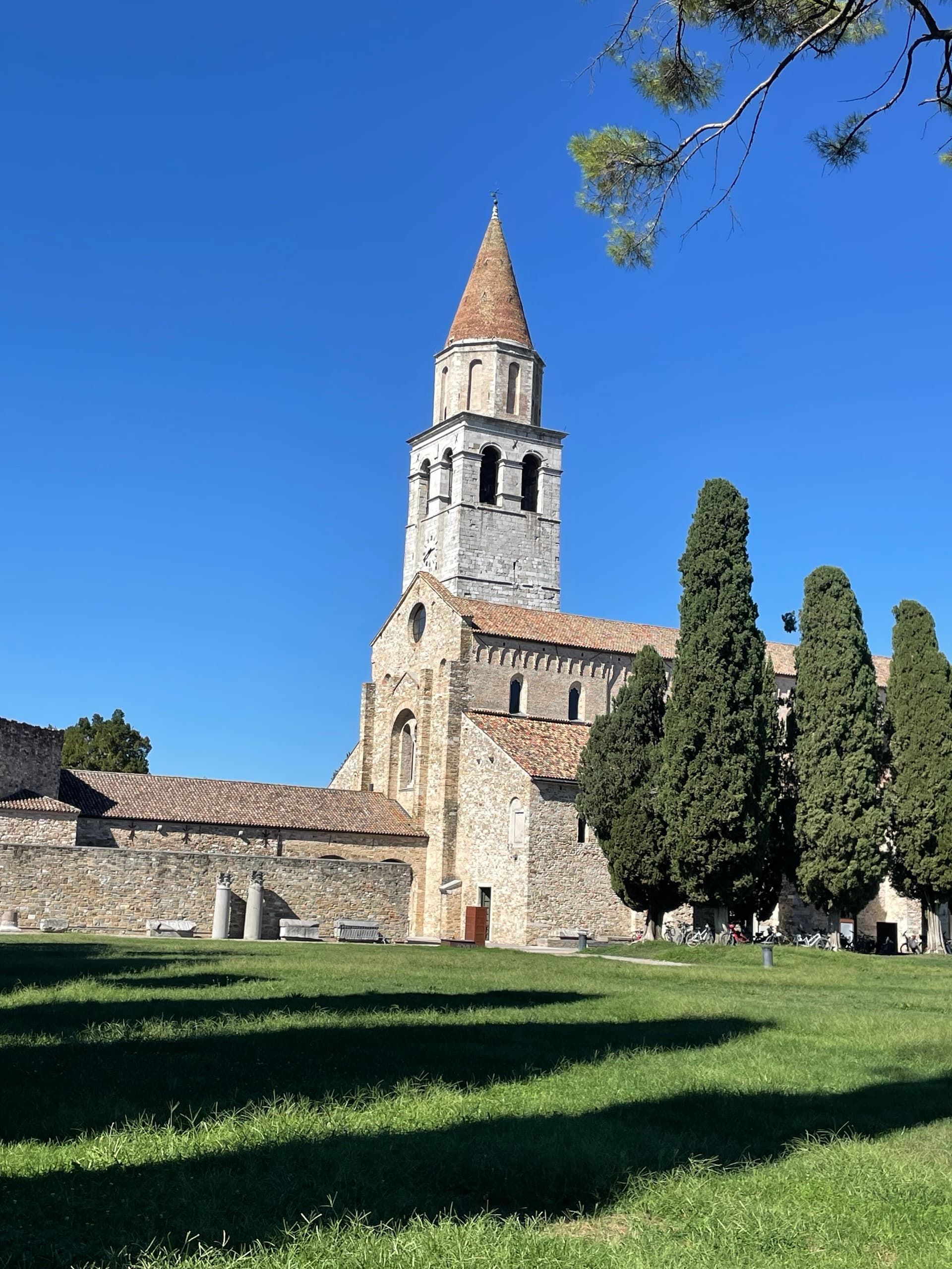 Aquileia Basilica and bell tower with stone architecture, cypress trees, and green lawn under a blue sky