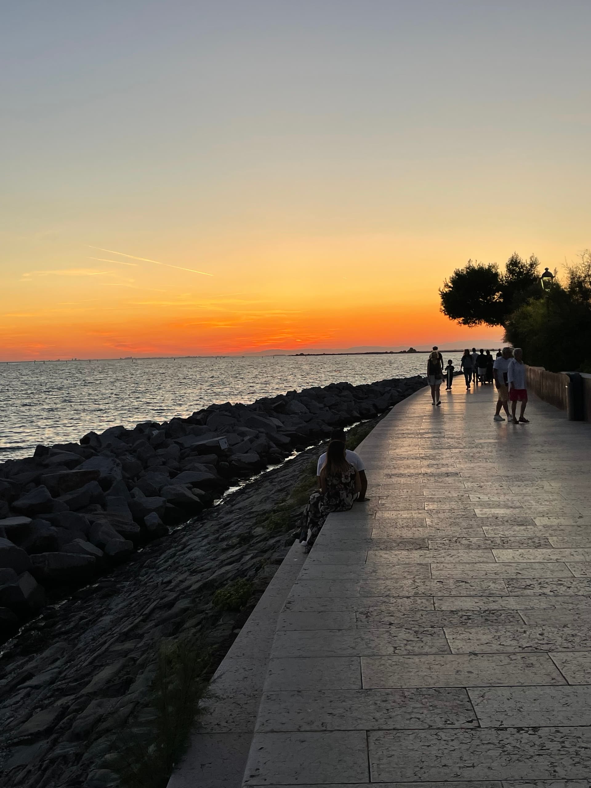 Grado seafront promenade at sunset with people walking and sitting by the rocky shore