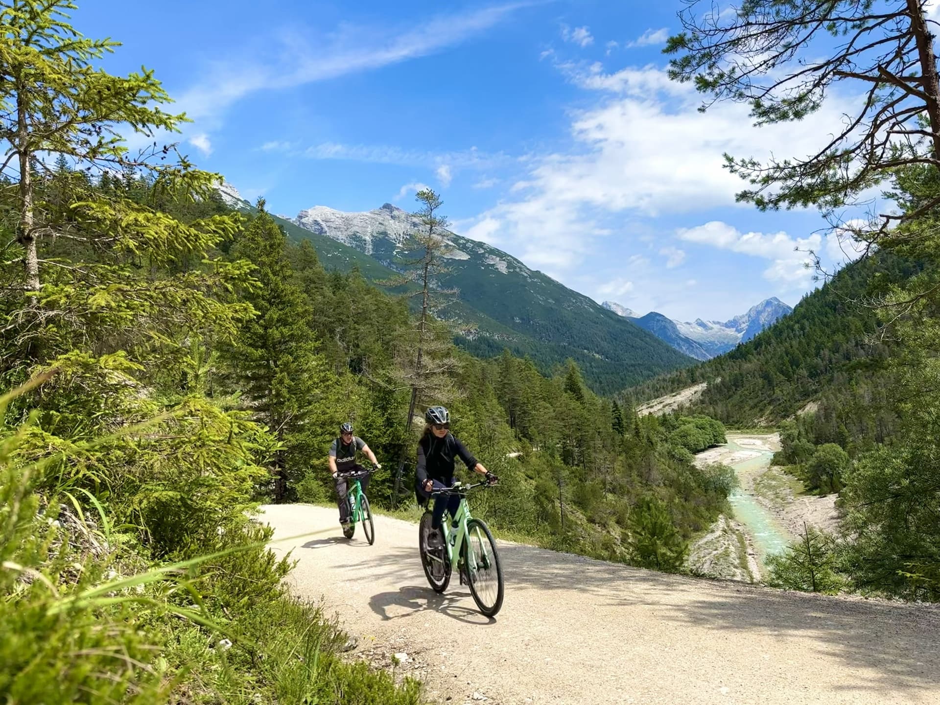 Cyclists descending on a gravel path above a mountain river valley with pine forests and peaks