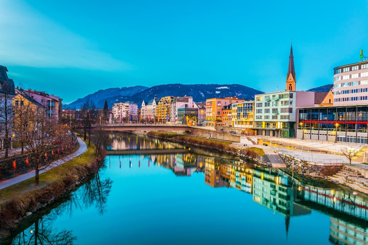 Riverfront buildings reflecting in bright blue water with mountains in Villach, Austria.