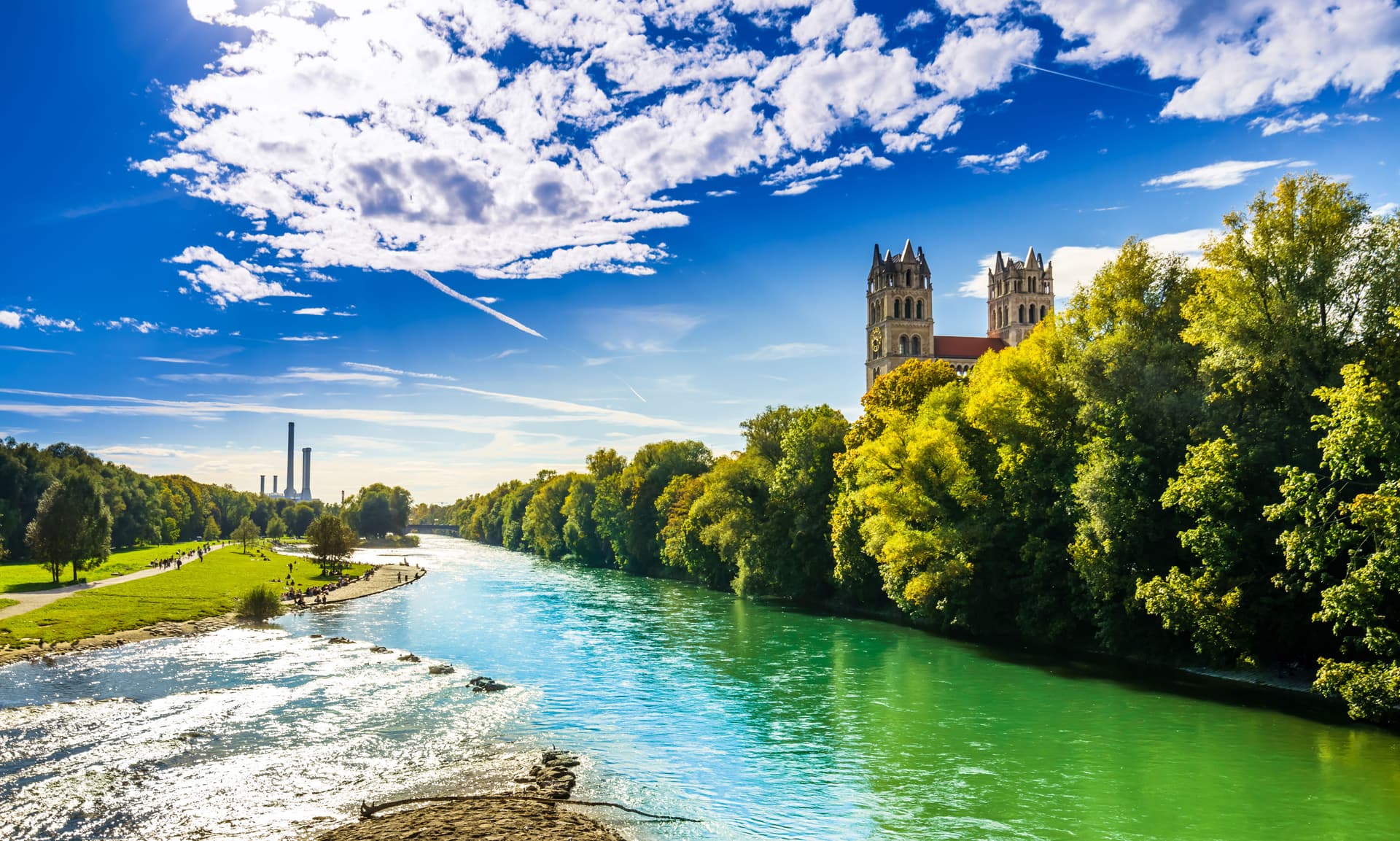 Isar River in Munich with autumn trees, St. Maximillian Church towers, and distant smokestacks.