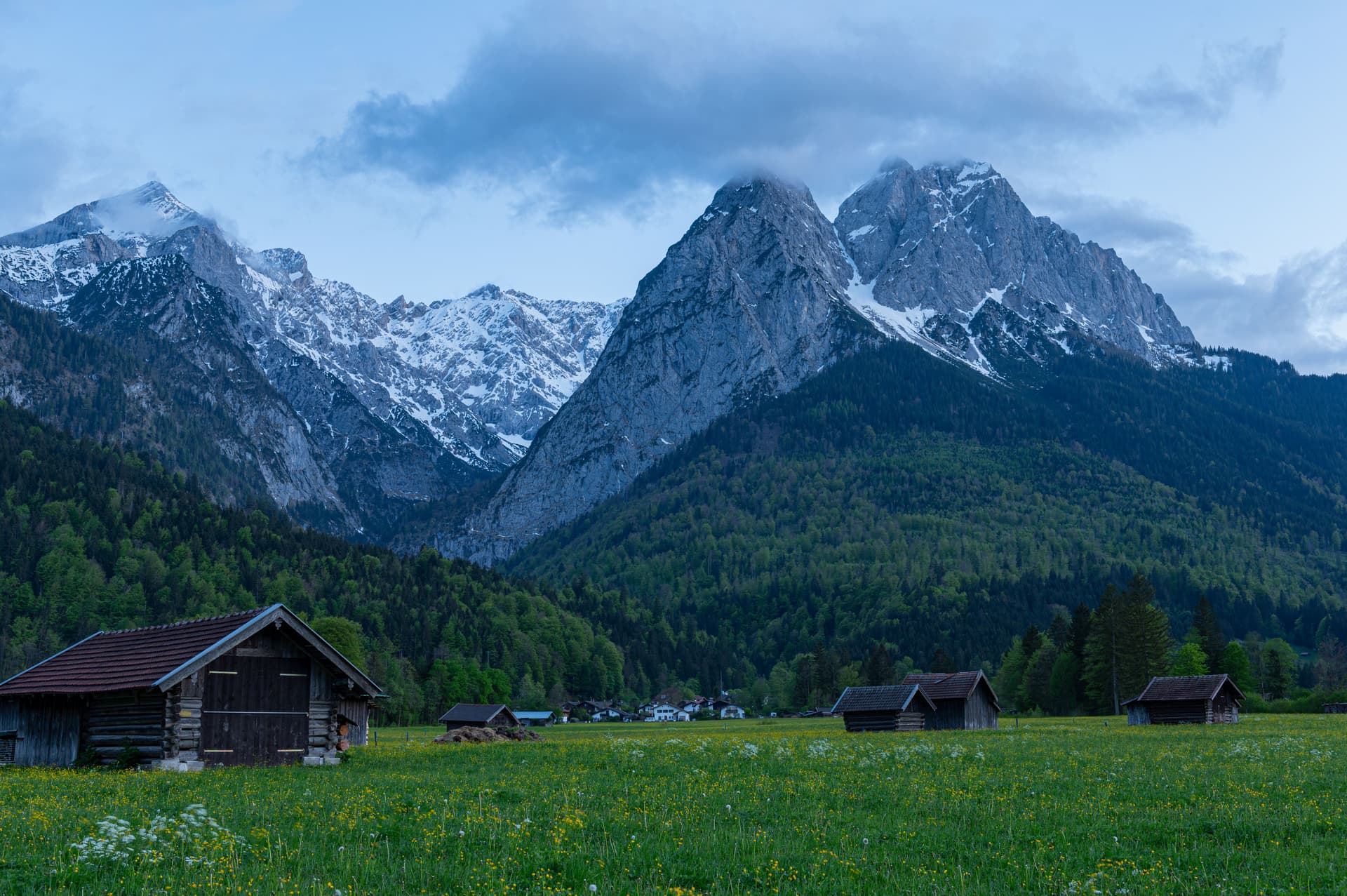 Wooden barns in a green meadow with yellow flowers below snowy alpine mountains.