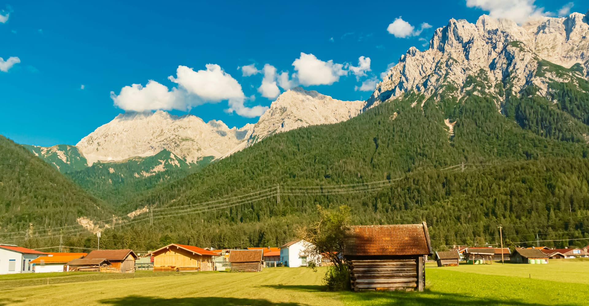 Alpine valley with wooden farm buildings, green fields, and towering rocky mountains under a blue sky.