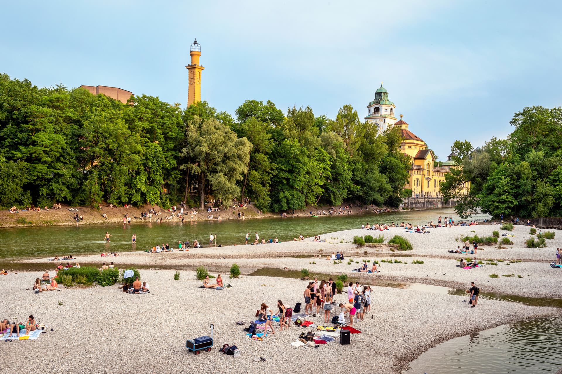 People sunbathing and swimming in the Isar River in Munich with historic buildings visible.