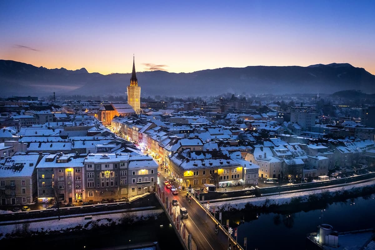 Snow-covered town of Villach at dusk with illuminated church tower and mountains in background.