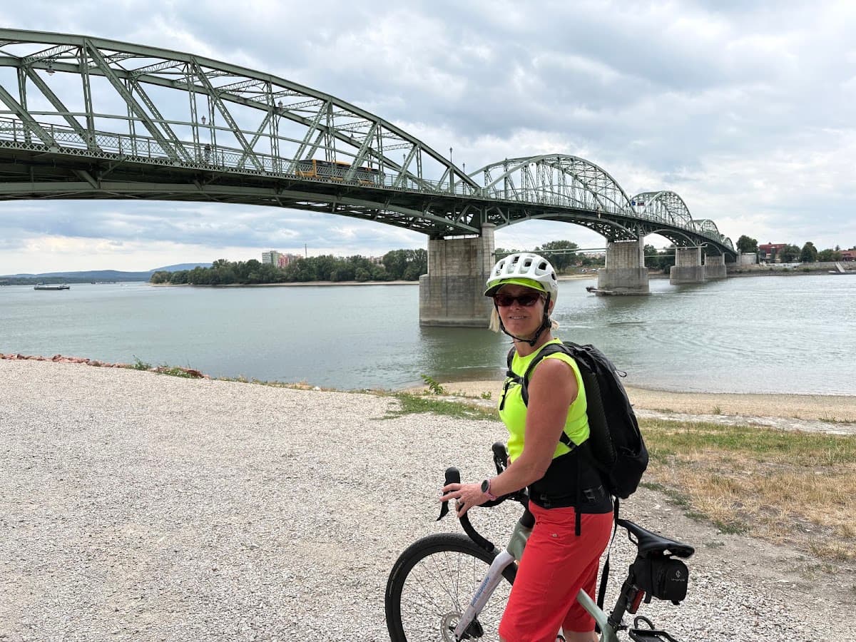 Cyclist with helmet and backpack standing with bicycle near large metal bridge over a river