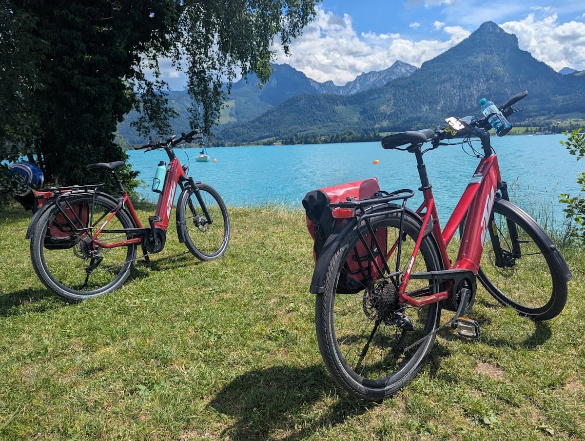 Two red bicycles parked on grass near an alpine lake with mountain peaks